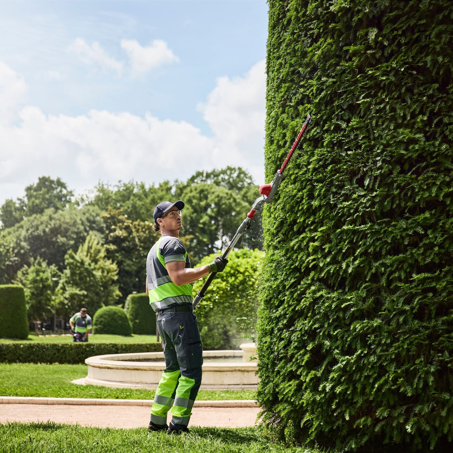 Person trimming a hedge using a hedge trimmer in a park setting.
