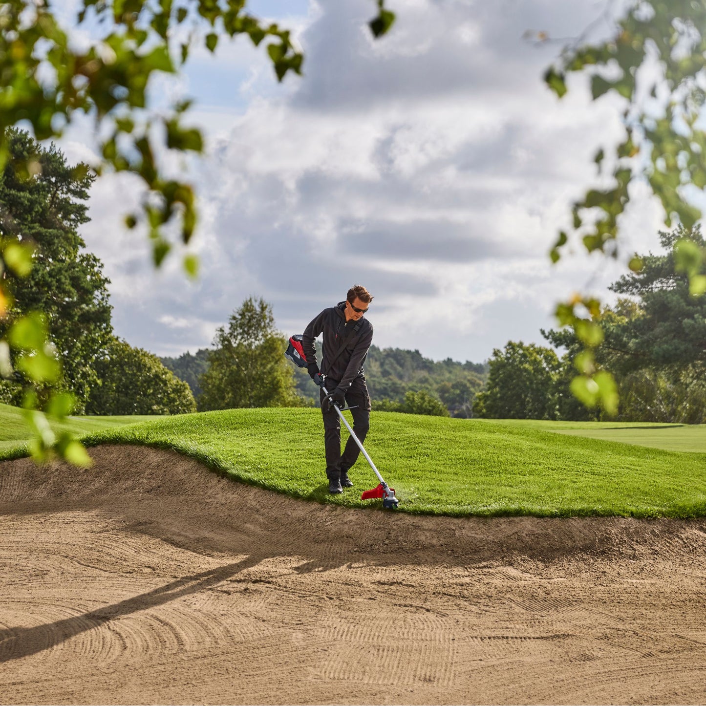 Person using a red and black strimmer on a golf course lawn edge with trees and sky in the background