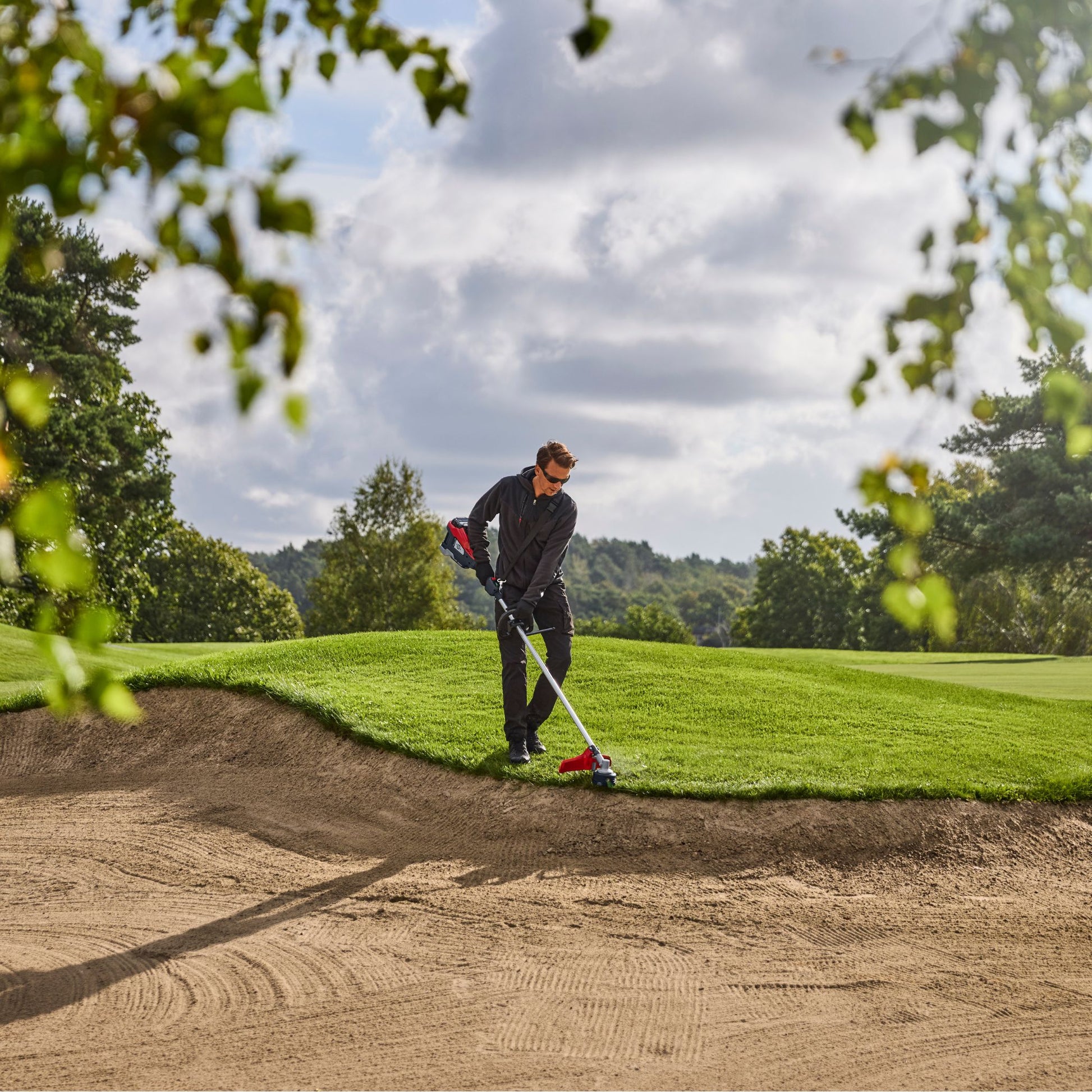 Person using a red and black strimmer on a golf course lawn edge with trees and sky in the background