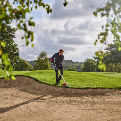 Person using a red and black strimmer on a golf course lawn edge with trees and sky in the background